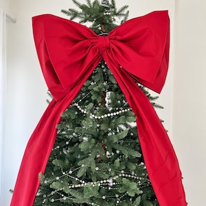 Könnte beinhalten: Eine große, leuchtend rote Stoffschleife an der Spitze eines grünen Weihnachtsbaums. Die Schleife hat zwei breite, strukturierte Schlaufen und zwei lange, herabhängende Bänder, die an den Seiten des mit silbernen Perlenketten geschmückten Baums verlaufen.