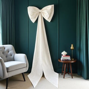 Pode incluir: Um grande laço de tecido em tom branco sujo com duas fitas longas e pontiagudas que pendem para baixo. O laço é feito de um material estruturado e liso, montado verticalmente para criar um elemento decorativo proeminente com dobras suaves.