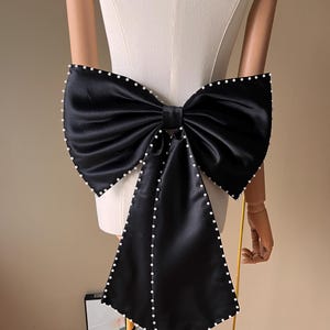 Könnte beinhalten: Eine große, schwarze Satinschleife mit Stoffkaskade. Die Schleife ist mit kleinen, weißen Perlen an den Rändern verziert, die einen Hauch von Eleganz verleihen. Die Schleife wird auf einer Schaufensterpuppe präsentiert.