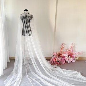 May include: A floor-length white bridal cape featuring a high collar with intricate white floral lace embroidery. The cape is constructed from sheer, lightweight tulle fabric that drapes from the shoulders down to the floor in a long train.