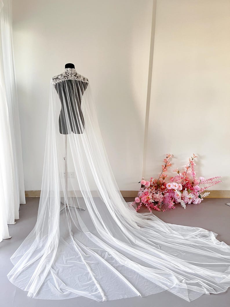 May include: A floor-length white bridal cape featuring a high collar with intricate white floral lace embroidery. The cape is constructed from sheer, lightweight tulle fabric that drapes from the shoulders down to the floor in a long train.