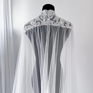 May include: A sheer white tulle cape featuring a high collar adorned with intricate silver-toned embroidery, beads, and sequins. The lightweight fabric drapes from the shoulders, creating a flowing silhouette with detailed floral-inspired embellishments.