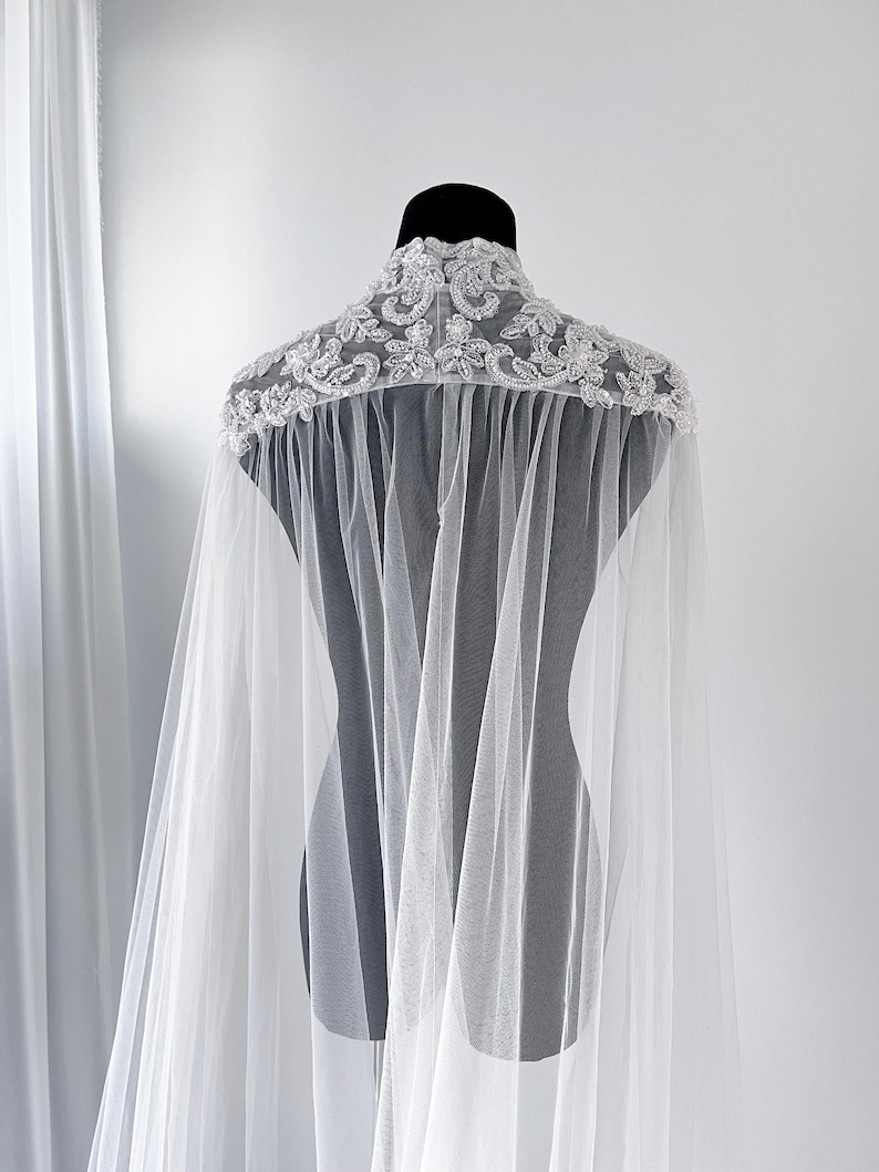 May include: A sheer white tulle cape featuring a high collar adorned with intricate silver-toned embroidery, beads, and sequins. The lightweight fabric drapes from the shoulders, creating a flowing silhouette with detailed floral-inspired embellishments.