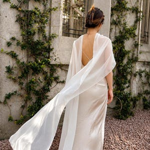 May include: A floor-length cream-coloured satin slip dress featuring a low back with thin crisscross straps and a long, flowing sheer white chiffon cape attached at the shoulders that drapes down the back, paired with beige heeled shoes.