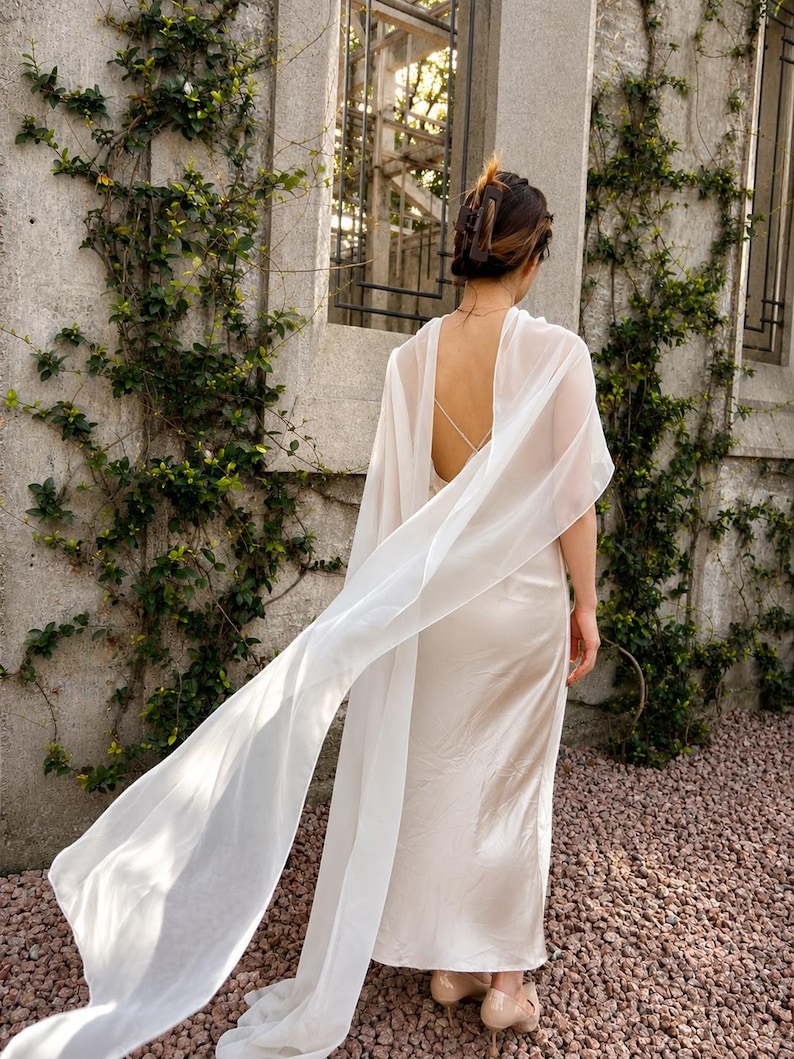 May include: A floor-length cream-coloured satin slip dress featuring a low back with thin crisscross straps and a long, flowing sheer white chiffon cape attached at the shoulders that drapes down the back, paired with beige heeled shoes.
