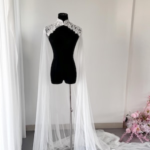 May include: A white floor-length bridal cape featuring a high collar adorned with intricate white floral lace embroidery. The cape is crafted from sheer, lightweight tulle fabric that drapes from the shoulders down to the floor in long panels.