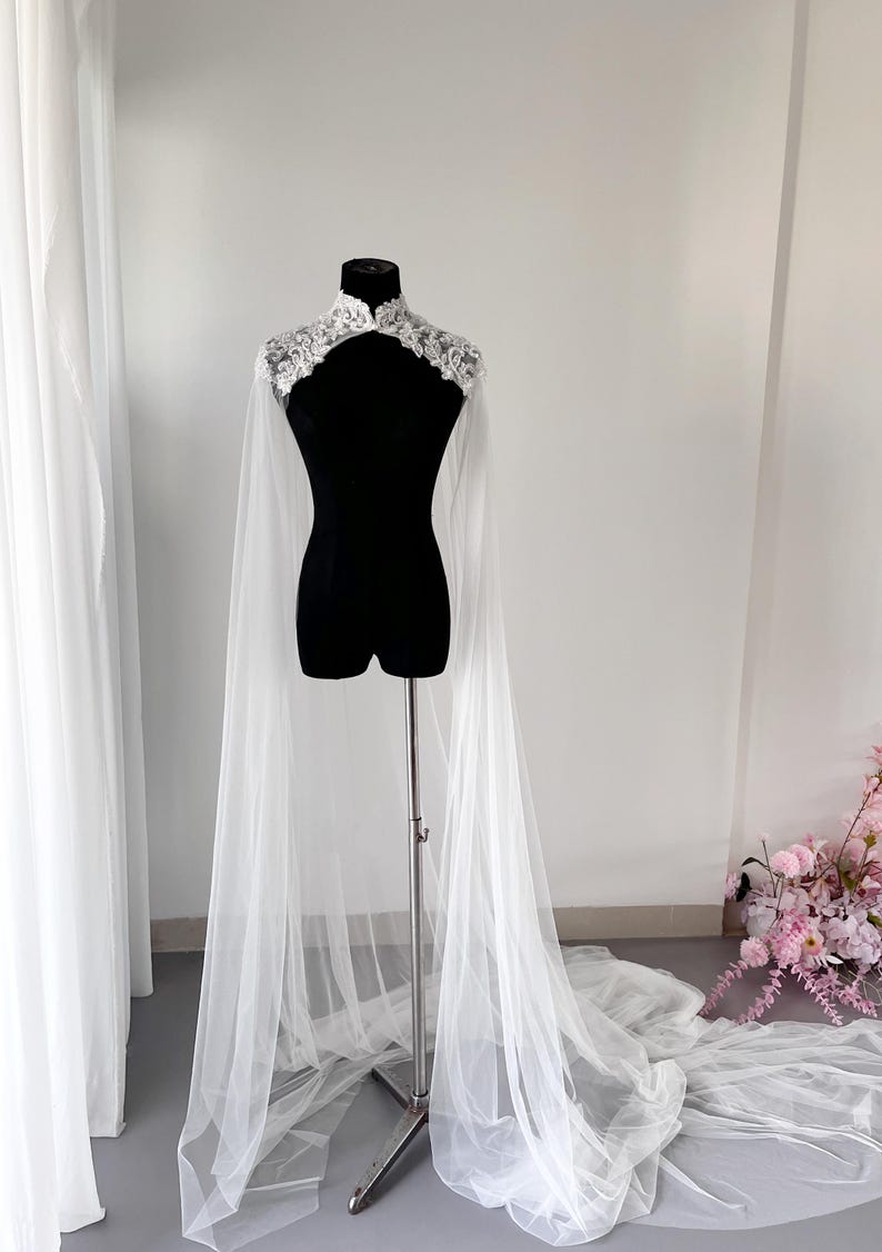 May include: A white floor-length bridal cape featuring a high collar adorned with intricate white floral lace embroidery. The cape is crafted from sheer, lightweight tulle fabric that drapes from the shoulders down to the floor in long panels.