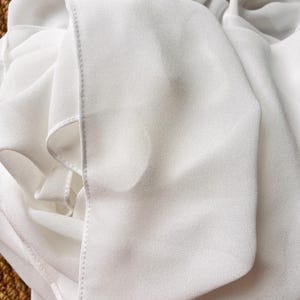 May include: A white garment made of a lightweight, semi-sheer fabric with a fine, crepe-like texture. The material is draped in soft folds, showcasing a clean, finished edge with visible stitching along the seam of the fabric panel.