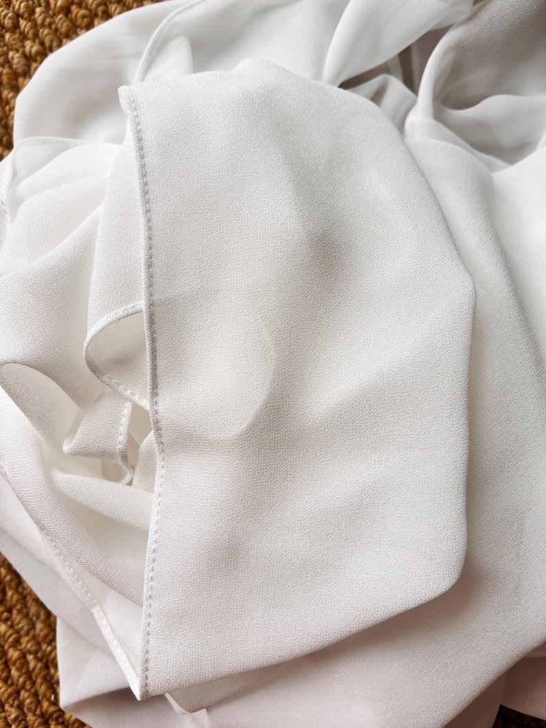 May include: A white garment made of a lightweight, semi-sheer fabric with a fine, crepe-like texture. The material is draped in soft folds, showcasing a clean, finished edge with visible stitching along the seam of the fabric panel.