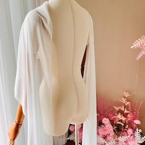 May include: A sheer white cape with long, flowing sleeves draped over a beige mannequin. The garment features a lightweight, translucent fabric that extends down from the shoulders, creating a soft, layered appearance on the torso and arms.