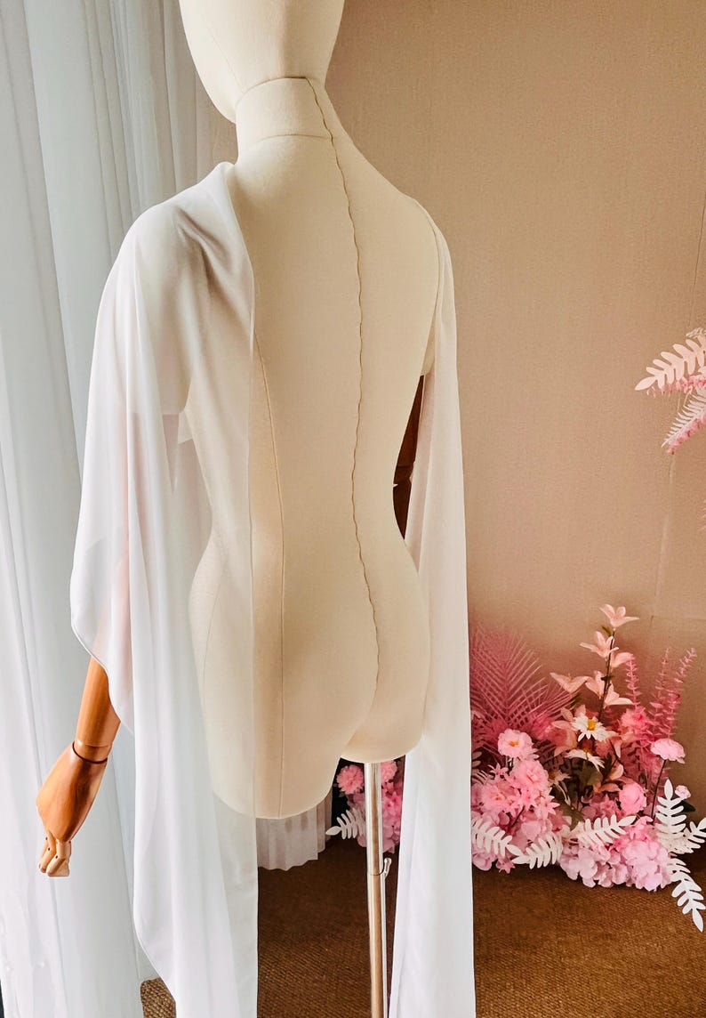 May include: A sheer white cape with long, flowing sleeves draped over a beige mannequin. The garment features a lightweight, translucent fabric that extends down from the shoulders, creating a soft, layered appearance on the torso and arms.