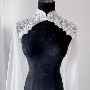 May include: A white bridal capelet featuring a high collar and intricate silver beadwork and embroidery across the shoulders. The garment is made of sheer, flowing fabric that drapes down from the embellished yoke, creating a cape-like effect.