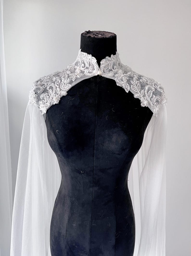 May include: A white bridal capelet featuring a high collar and intricate silver beadwork and embroidery across the shoulders. The garment is made of sheer, flowing fabric that drapes down from the embellished yoke, creating a cape-like effect.