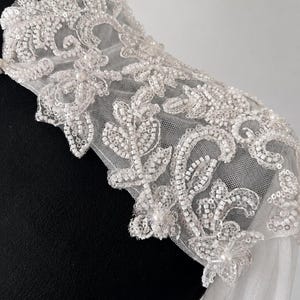 May include: A white sheer tulle shoulder piece featuring intricate embroidery with clear sequins, glass beads, and small white pearls. The floral-inspired pattern covers the high collar and extends over the shoulder, creating a textured finish.