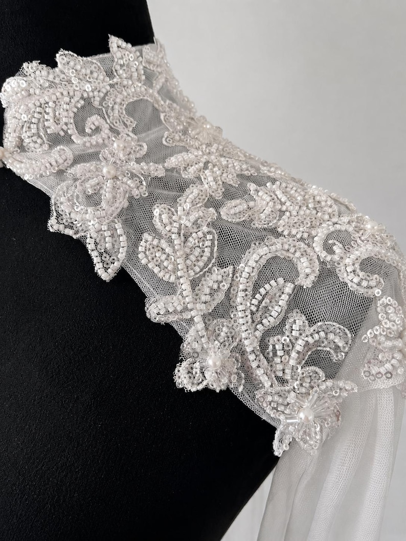 May include: A white sheer tulle shoulder piece featuring intricate embroidery with clear sequins, glass beads, and small white pearls. The floral-inspired pattern covers the high collar and extends over the shoulder, creating a textured finish.