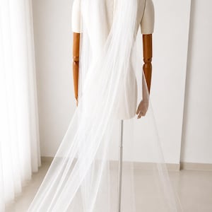 May include: A long, sheer white bridal veil draped over a mannequin. The veil is made of a delicate, flowing fabric and is displayed against a white background. The mannequin has wooden arms and is mounted on a metal stand.