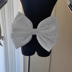 May include: A large white lace bow on a black mannequin. The bow is tied in a large, dramatic knot.