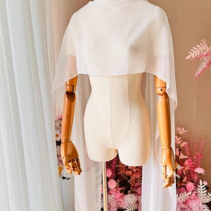 May include: A sheer white chiffon capelet draped over a mannequin. The garment features a high neckline and long, flowing panels that extend down from the shoulders, creating a lightweight, semi-transparent layer with clean, finished edges.
