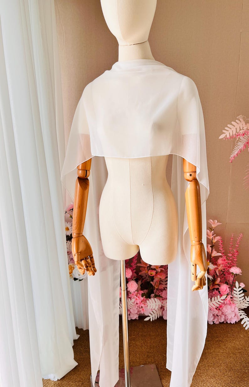 May include: A sheer white chiffon capelet draped over a mannequin. The garment features a high neckline and long, flowing panels that extend down from the shoulders, creating a lightweight, semi-transparent layer with clean, finished edges.