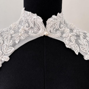 May include: A white lace collar featuring intricate floral embroidery, clear sequins, and small white beads. The collar has a high neckline with a single pearl button closure at the center front, crafted from sheer mesh fabric with scalloped edges.