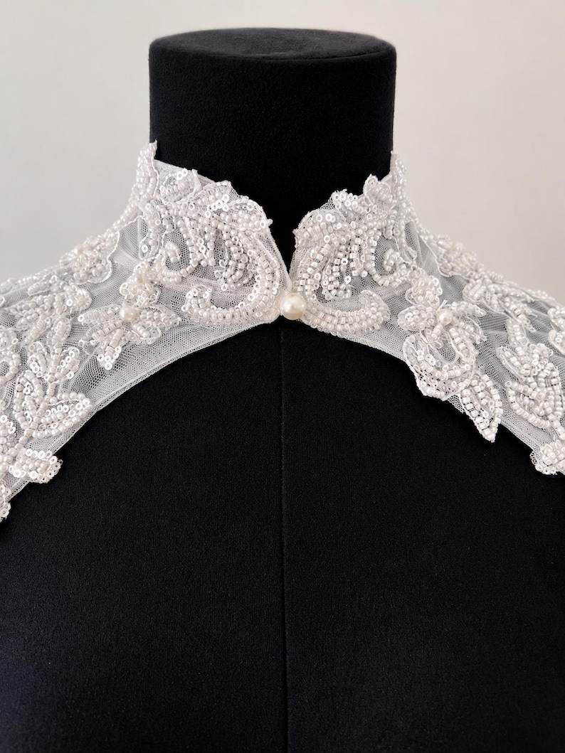 May include: A white lace collar featuring intricate floral embroidery, clear sequins, and small white beads. The collar has a high neckline with a single pearl button closure at the center front, crafted from sheer mesh fabric with scalloped edges.