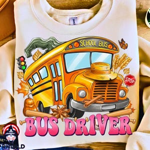 Op de afbeelding: Crèmekleurige sweatshirt met een kleurrijke schoolbusafbeelding en de roze tekst "BUS DRIVER". Het ontwerp bevat herfstthema-elementen zoals bladeren, een pompoen en een stopbord, wat een seizoensgebonden en thematische aantrekkingskracht creëert.
