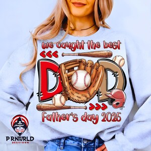 Op de afbeelding: Lichtgrijze sweatshirt met een honkbalthema. Het ontwerp bevat het woord "DAD" in rood en bruin, honkballen, knuppels, een handschoen en een helm. Tekst luidt "We caught the best Father's day 2025."