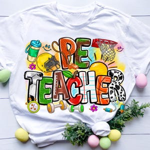 Può includere: T-shirt bianca con un design grafico colorato. Il design presenta le parole "PE TEACHER" in vari font e colori, insieme a attrezzature sportive come un pallone da basket, football americano e manubri. Elementi aggiuntivi includono una borraccia, una mela e fiori.