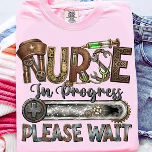 May include: A pink t-shirt with a steampunk-style design. The design includes the word "NURSE" with gears and medical tools, the phrase "In Progress," and "PLEASE WAIT." The shirt has a crew neck and a "Comfort Colors" tag.