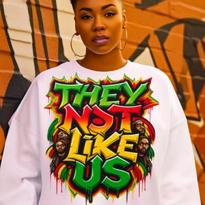 May include: White sweatshirt featuring the text "THEY NOT LIKE US" in green, red, and yellow. The design includes illustrations of people with dreadlocks. The sweatshirt has a crew neck.