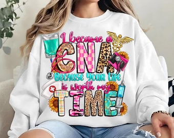 May include: White sweatshirt with a colorful graphic design that says "I became a CNA because your life is worth my time!" The graphic includes a stethoscope, a heart, a syringe, a bandage, and a sunflower.