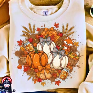 May include: Cream-colored sweatshirt featuring a fall-themed graphic. The design includes orange pumpkins, baseballs with bows, wheat stalks, red apples, and a fox wearing a scarf. A basket of pumpkins and jars of spices complete the autumn scene.