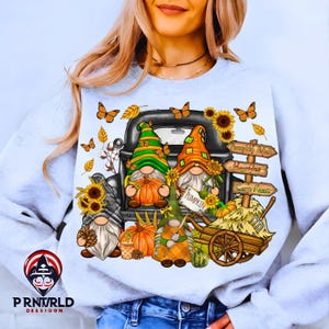 Puede incluir: Una sudadera gris claro con un gráfico temático de otoño. El diseño incluye tres gnomos en la caja de una camioneta negra, calabazas, girasoles, mariposas y letreros que dicen "Pumpkin Patch", "Hayrides" y "Corn Maze".