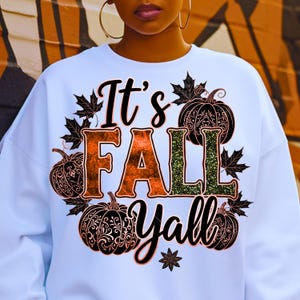 May include: White sweatshirt with the text "It's FALL Y'all" in orange and green, surrounded by black pumpkins and maple leaves. The design has a fall theme.