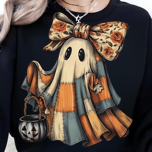 May include: Black sweatshirt featuring a whimsical ghost design. The ghost is adorned with a patchwork quilt-like appearance in orange, blue, and cream colors. It has a floral bow and carries a jack-o'-lantern bucket, perfect for Halloween.