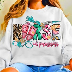Puede incluir: Sudadera blanca con un diseño gráfico colorido. La palabra "NURSE" está escrita en varios patrones, incluyendo estampado de leopardo y cuadros. También se presentan suministros médicos y la frase "in progress".