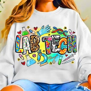 以下が含まれることがあります： カラフルな模様の文字で「LAB TECH」と書かれた白いスウェットシャツ。ビーカー、顕微鏡、DNA鎖など、実験室関連のイラストがデザインされています。カジュアルで快適なスタイルです。
