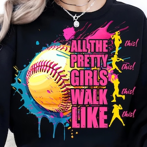 May include: Black sweatshirt with a colorful graphic design. The design features a softball with pink and yellow accents, and the text "ALL THE PRETTY GIRLS WALK LIKE THIS!" in pink. Silhouettes of softball players are also included.
