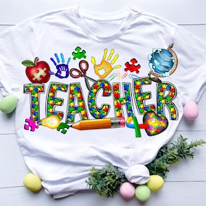 May include: White t-shirt with a colorful graphic design that says "TEACHER" in puzzle pieces. The design also includes a lightbulb, pencil, scissors, a globe, a heart, and handprints.