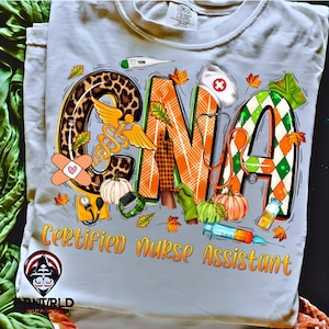 May include: Light blue t-shirt with a colorful CNA design. The design includes the letters "CNA" with leopard print, plaid, and diamond patterns. The words "Certified Nurse Assistant" are in orange. Pumpkins, leaves, and medical tools are also featured.