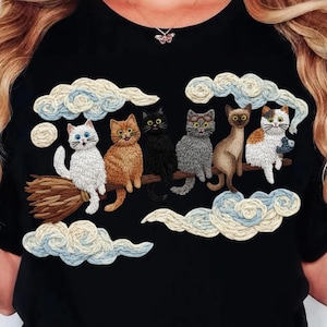 Könnte beinhalten: Schwarzes T-Shirt mit einem gestickten Design von sechs Katzen, die auf einem Besen durch einen bewölkten Himmel fliegen. Die Katzen haben verschiedene Farben, darunter Weiß, Orange, Schwarz und Grau. Die Wolken sind weiß und blau.