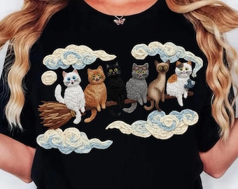 Pode incluir: T-shirt preto com um desenho bordado de seis gatos numa vassoura a voar num céu nublado. Os gatos são de várias cores, incluindo branco, laranja, preto e cinzento. As nuvens são brancas e azuis.