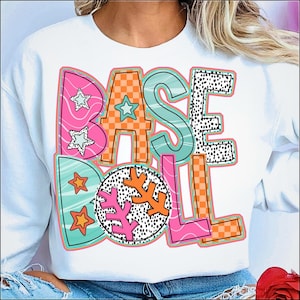 May include: White sweatshirt with the word "BASEBALL" in colourful, playful lettering. The letters feature various patterns, including stars, checkerboard, and polka dots, with a baseball-themed design.