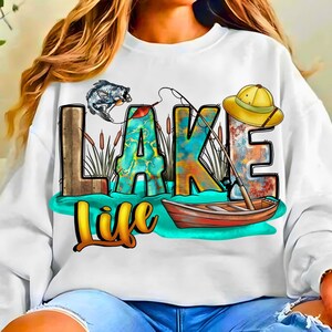 May include: White sweatshirt with a colorful graphic design featuring the words "LAKE LIFE". The design includes a fishing rod, a fish, a boat, and a yellow hat. The letters are in a variety of colors, including teal, orange, and brown.