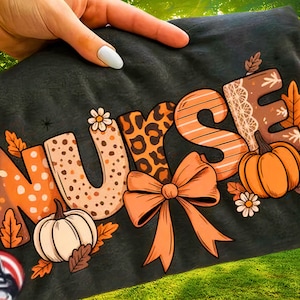 May include: Dark grey t-shirt featuring the word "NURSE" in a seasonal design. The letters are decorated with pumpkins, leaves, and a bow. The "U" has a daisy, and the "R" has a leopard print.