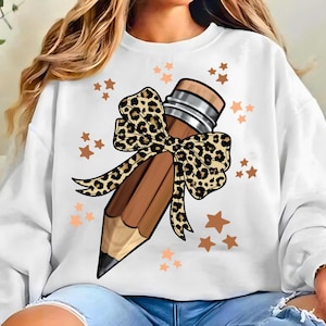 May include: White sweatshirt featuring a large brown pencil design with a leopard print bow. The pencil is accented with silver bands and a black tip. Scattered around the pencil are small, copper-colored star shapes.