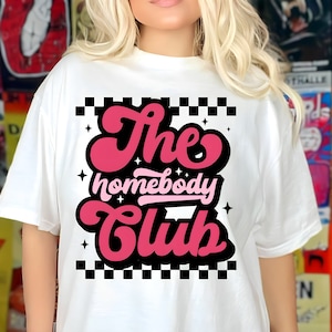 Puede incluir: Camiseta blanca con el texto "The Homebody Club" en un diseño retro en rosa y rojo. El diseño está enmarcado por un patrón de cuadros blancos y negros. La camiseta es de corte casual.