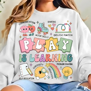 May include: White sweatshirt with a colorful graphic that reads "PLAY IS LEARNING." The design includes illustrations of a brain, apple, roller skate, hearts, and other educational symbols. Text includes "COMMUNICATION", "MOTOR SKILLS", and "SELF REGULATION".