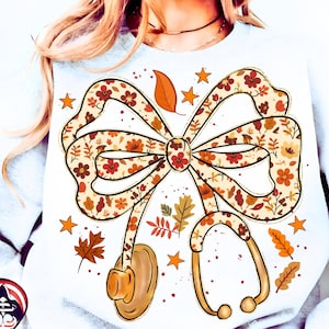 Puede incluir: Sudadera blanca con un gran diseño de lazo con un estampado floral en colores otoñales. Un estetoscopio está unido al lazo, con hojas y estrellas dispersas. El diseño tiene un tema otoñal o médico.