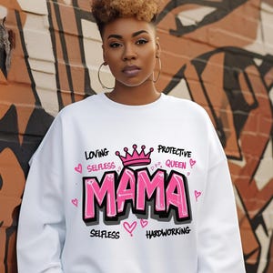 May include: White sweatshirt with a pink and black graphic design that says "MAMA" with a crown above the word and the words "Loving, Selfless, Protective Queen, Selfless, Hardworking" around the word "MAMA".